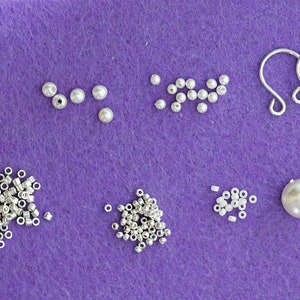 Może przedstawiać: Zestaw białych pereł, srebrnych k&oacute;łek łączących i srebrnych haczyk&oacute;w do kolczyk&oacute;w na fioletowym filcu.