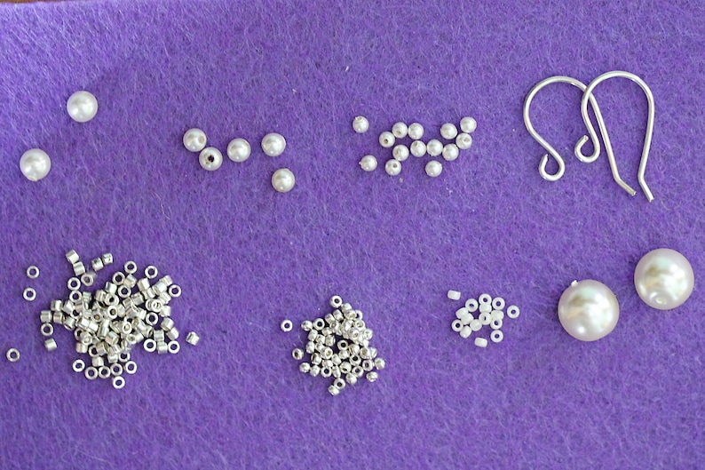 Może przedstawiać: Zestaw białych pereł, srebrnych k&oacute;łek łączących i srebrnych haczyk&oacute;w do kolczyk&oacute;w na fioletowym filcu.