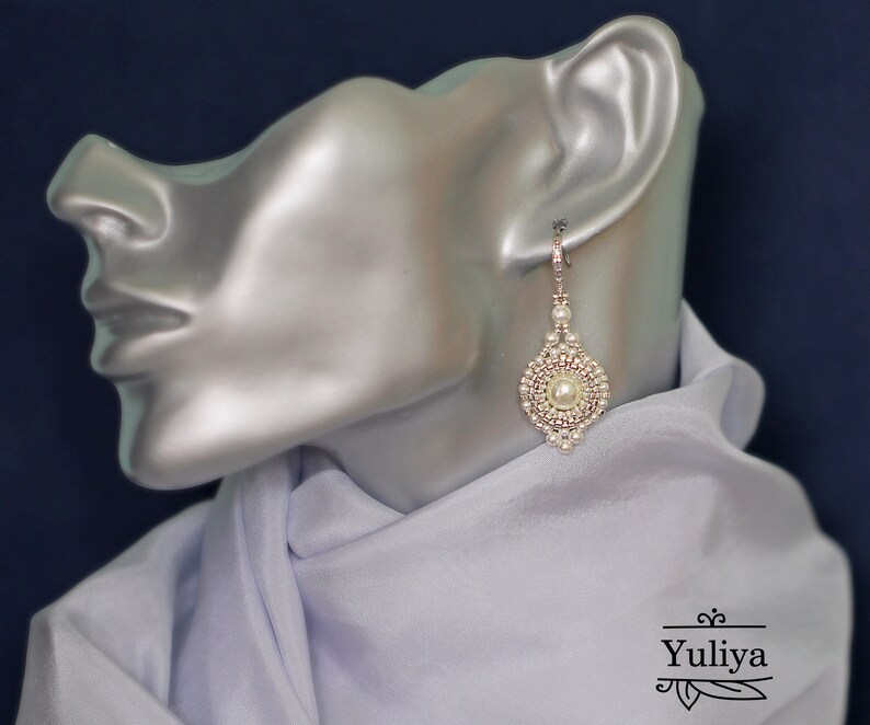 Może przedstawiać: Para białych perełkowych kolczyk&oacute;w wiszących z srebrnym zapięciem. Kolczyki są koralikowe i mają kształt łzy.