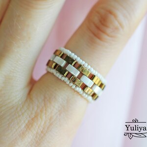 Peut inclure: Une bague en perles dor&eacute;es et blanches avec un motif g&eacute;om&eacute;trique. La bague est sur un doigt.