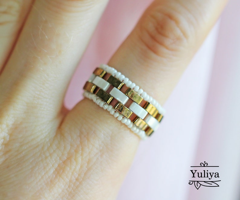 Peut inclure: Une bague en perles dor&eacute;es et blanches avec un motif g&eacute;om&eacute;trique. La bague est sur un doigt.