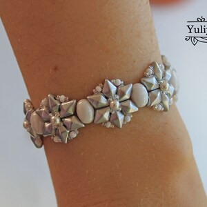 Puede incluir: Un brazalete de cuentas plateadas y blancas con un dise&ntilde;o de flores. El brazalete est&aacute; en la mu&ntilde;eca de una persona.