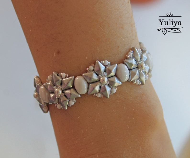 Puede incluir: Un brazalete de cuentas plateadas y blancas con un dise&ntilde;o de flores. El brazalete est&aacute; en la mu&ntilde;eca de una persona.
