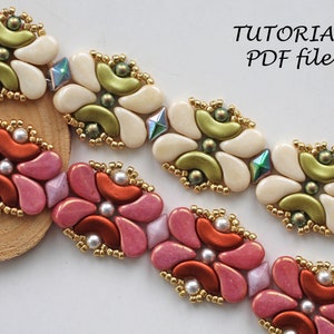 Op de afbeelding: Twee kralenarmbanden met een bloemenmotief. De bovenste armband is wit met groene accenten en de onderste armband is roze met oranje accenten. Beide armbanden hebben gouden kralen.