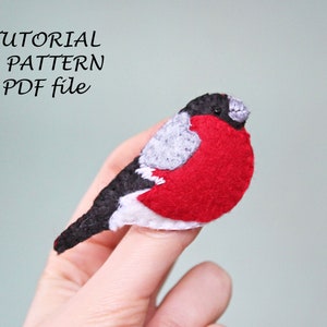 May include: A felt bird with a red breast, grey wings, and a black head. The bird is being held by a finger. The text "TUTORIAL & PATTERN PDF file" is written above the bird.