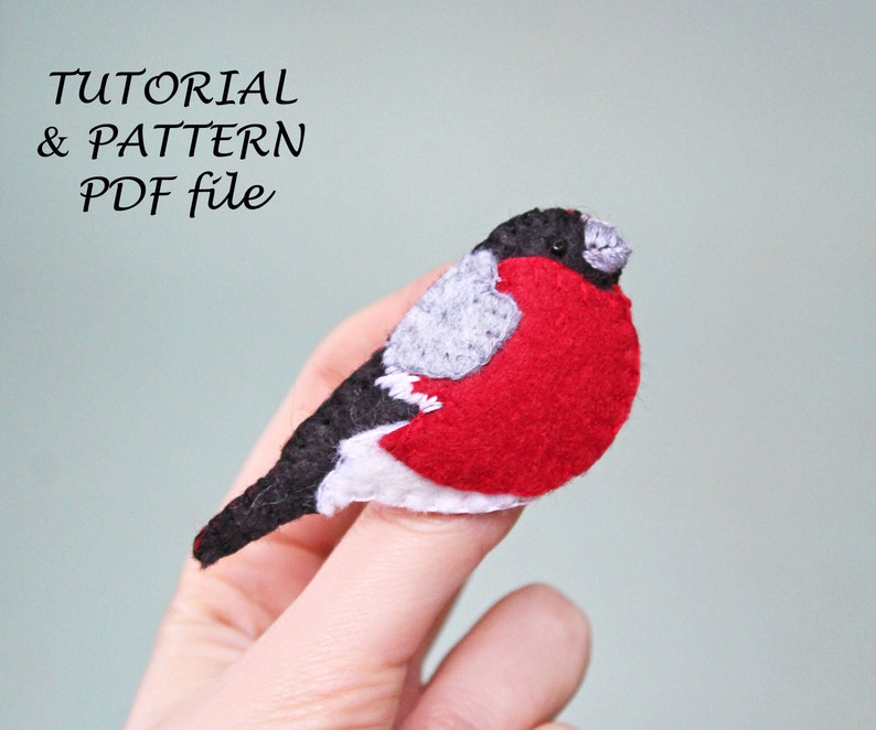May include: A felt bird with a red breast, grey wings, and a black head. The bird is being held by a finger. The text "TUTORIAL & PATTERN PDF file" is written above the bird.