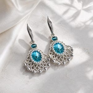 Può includere: Un paio di orecchini pendenti con un grande cristallo centrale ovale di colore blu brillante circondato da intricate perline argentate. Ogni orecchino include un cristallo blu rotondo più piccolo e un perno in metallo argentato liscio.