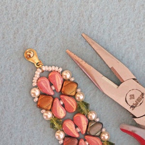 May include: Close-up of a beaded bracelet with pink, gold, green, and white beads. The bracelet is being held by a pair of pliers with a red handle.