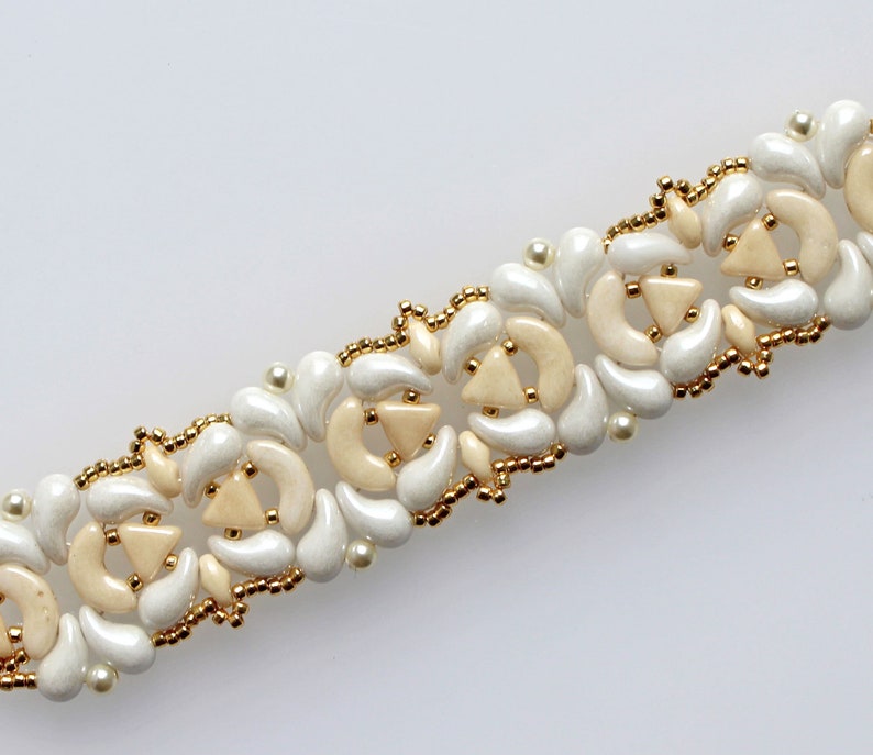 Op de afbeelding: Een witte en cr&egrave;mekleurige kralenarmband met een delicaat bloemenpatroon. De armband is gemaakt van kleine, ronde kralen en heeft een herhalend patroon van bloemen.