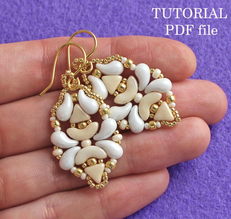 Op de afbeelding: Een paar gouden oorbellen met witte en cr&egrave;mekleurige kralen in een geometrisch patroon. De oorbellen worden in een hand tegen een paarse achtergrond gehouden. De tekst "TUTORIAL PDF file" is zichtbaar in de rechterbovenhoek van de afbeelding.