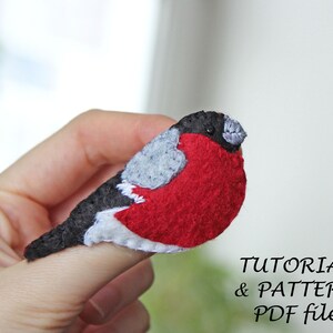 May include: A felt bird pin with a black head, grey wings, and a red body. The pin is held on a finger. The text "TUTORIAL & PATTERN PDF file" is below the bird.