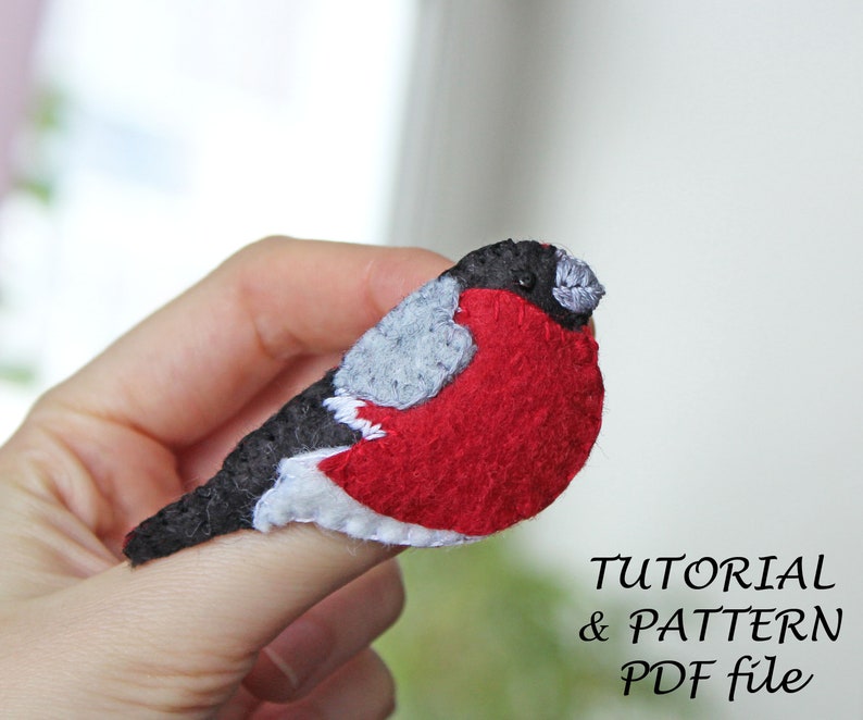 May include: A felt bird pin with a black head, grey wings, and a red body. The pin is held on a finger. The text "TUTORIAL & PATTERN PDF file" is below the bird.