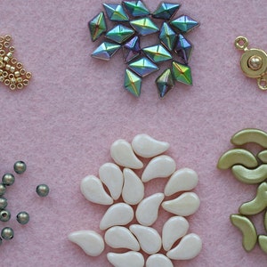 Op de afbeelding: Een assortiment kralen in verschillende vormen, kleuren en maten, waaronder gouden zaadkralen, iriserende groene gefacetteerde kralen, witte traanvormige kralen, groene halvemaanvormige kralen en grijze ronde kralen. Ook inbegrepen is een gouden metalen sluiting met een bal in het midden.