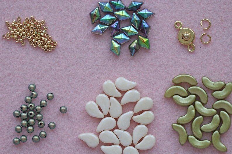 Op de afbeelding: Een assortiment kralen in verschillende vormen, kleuren en maten, waaronder gouden zaadkralen, iriserende groene gefacetteerde kralen, witte traanvormige kralen, groene halvemaanvormige kralen en grijze ronde kralen. Ook inbegrepen is een gouden metalen sluiting met een bal in het midden.
