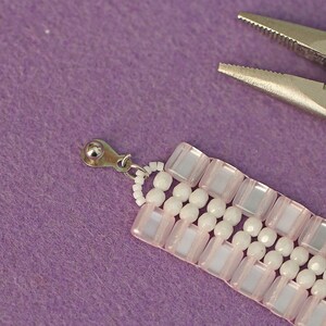 May include: A close-up of a pink and white beaded bracelet with a silver clasp. The bracelet is being held by a pair of pliers.