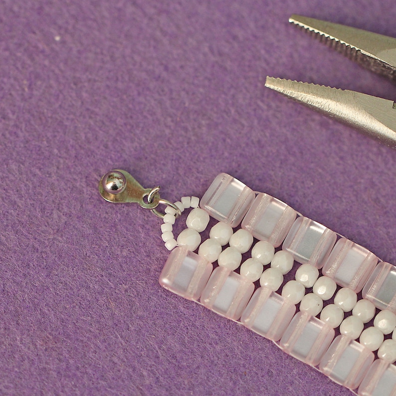 May include: A close-up of a pink and white beaded bracelet with a silver clasp. The bracelet is being held by a pair of pliers.