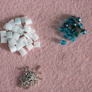 May include: Three piles of beads on a pink surface. One pile is white, one is silver, and one is teal.