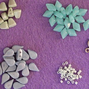 Peut inclure: Divers &eacute;l&eacute;ments pour la cr&eacute;ation de bijoux, comprenant des perles en forme de goutte et de losange bleu clair, de petites perles tubulaires blanches, un fermoir argent&eacute; et des anneaux. Le tout est dispos&eacute; sur un fond en feutre violet.