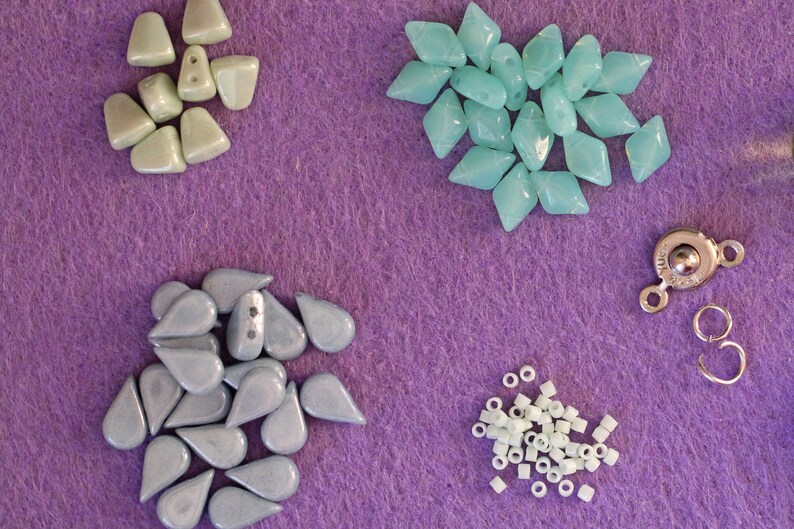 Peut inclure: Divers &eacute;l&eacute;ments pour la cr&eacute;ation de bijoux, comprenant des perles en forme de goutte et de losange bleu clair, de petites perles tubulaires blanches, un fermoir argent&eacute; et des anneaux. Le tout est dispos&eacute; sur un fond en feutre violet.