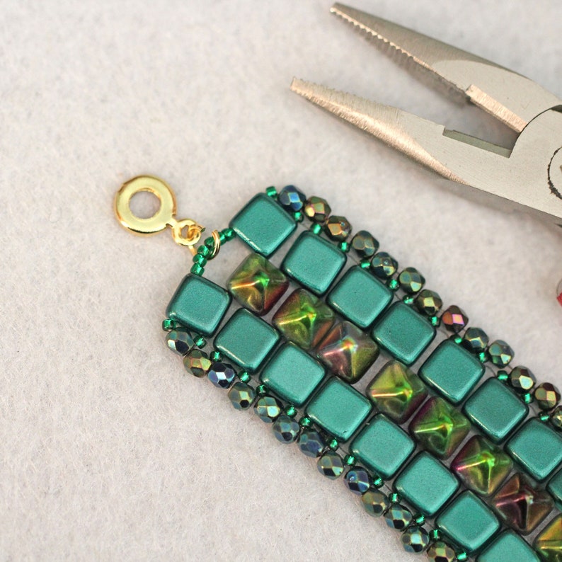 May include: A green and gold beaded bracelet with a clasp. The bracelet is made with square and round beads.