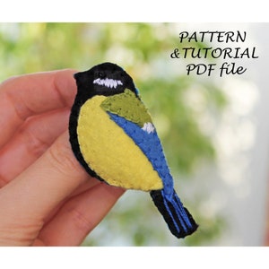May include: A felt bluebird with black, white, and green accents. The bird is a craft project and is being held in a hand. The image includes the text "PATTERN & TUTORIAL PDF file".