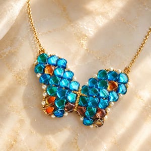 May include: A butterfly-shaped pendant necklace featuring iridescent blue, teal, and amber glass scales arranged in a wing pattern, accented with small white pearls along the edges and a gold-toned metal chain and central body detail.