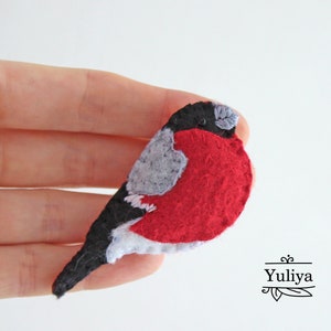 May include: A felt bird brooch with a red body, grey wings, and black accents. The bird is perched on a hand.