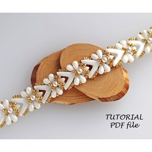 Könnte beinhalten: Weißes und goldenes Perlenarmband mit Chevron-Muster und Blumenakzenten. Das Armband liegt auf einer Holzoberfläche. TUTORIAL PDF file.