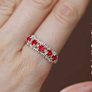 May include: A silver ring with red gemstones set in a beaded band. The ring is worn on a person's finger.
