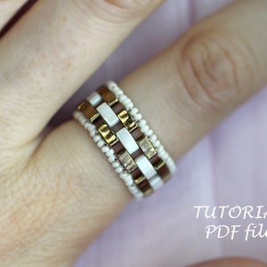 Peut inclure: Gros plan d'une main portant une bague en perles avec des perles carr&eacute;es blanches et dor&eacute;es. La bague est sur un doigt avec un fond rose clair. Le texte "TUTORIAL PDF file" est visible dans le coin inf&eacute;rieur droit de l'image.