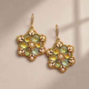 Puede incluir: Un par de pendientes colgantes con un diseño floral hexagonal, elaborados con cuentas doradas, pequeñas perlas blancas y cabujones de vidrio translúcido con textura en tonos verde y amarillo. Cada pendiente cuelga de un gancho dorado con cristales.