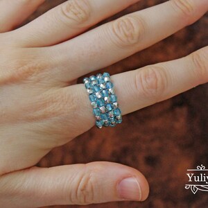 May include: A close-up of a hand wearing a ring made of multiple rows of small, light blue and silver beads.