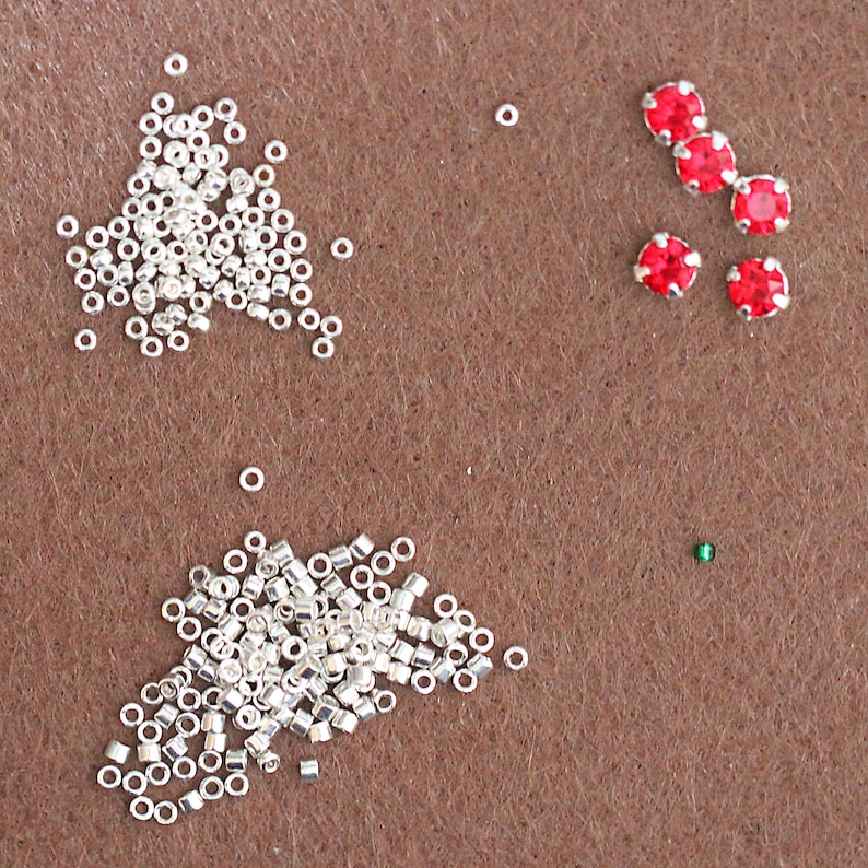May include: Two piles of silver jump rings and a scattering of red and green rhinestones on a brown surface.