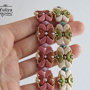 Op de afbeelding: Twee kralenarmbanden met een bloemenpatroon. De ene armband is gemaakt met roze, rode en gouden kralen. De andere armband is gemaakt met witte, groene en gouden kralen.