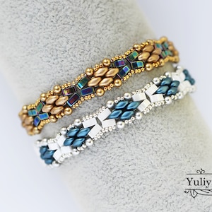 Op de afbeelding: Twee kralenarmbanden, een met gouden en regenboogkleurige blauwe kralen, de andere met zilveren en blauwgroene kralen. De armbanden liggen op een grijs oppervlak.