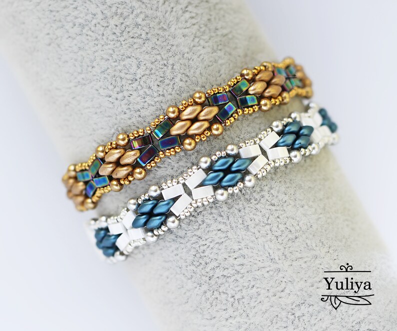 Op de afbeelding: Twee kralenarmbanden, een met gouden en regenboogkleurige blauwe kralen, de andere met zilveren en blauwgroene kralen. De armbanden liggen op een grijs oppervlak.