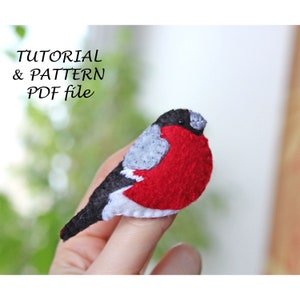May include: A felt bird brooch with a red body, grey wings, and black head. The bird is perched on a finger.