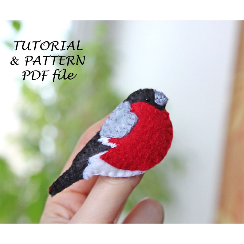 May include: A felt bird brooch with a red body, grey wings, and black head. The bird is perched on a finger.
