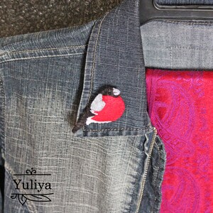 May include: A felt bird brooch in the shape of a red-breasted robin. The bird is perched on a denim jacket with a red scarf.