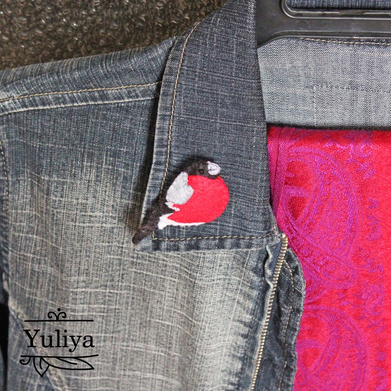 May include: A felt bird brooch in the shape of a red-breasted robin. The bird is perched on a denim jacket with a red scarf.