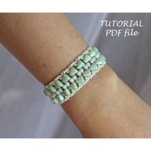 Op de afbeelding: Een groene en witte kralen armband met een geometrisch patroon. De armband zit om de pols van een persoon. De afbeelding bevat de tekst "TUTORIAL PDF file".