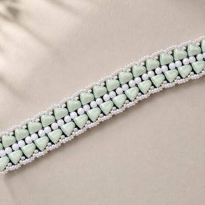 May include: A beaded bracelet featuring a central row of triangular, pale mint green glass beads flanked by two rows of small, round white seed beads. The design is symmetrical, with the mint green beads alternating in orientation along the band.
