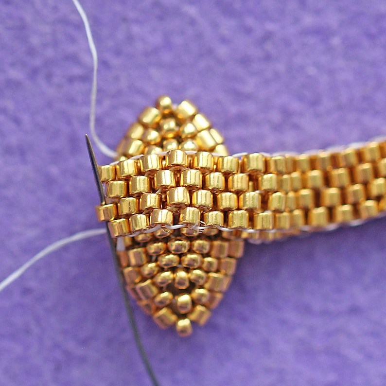 K&ouml;nnte beinhalten: Nahaufnahme eines goldenen Schmuckst&uuml;cks aus Perlen, durch das eine Nadel mit Faden gef&uuml;hrt wird. Die Perlen sind in einem geometrischen Muster angeordnet.