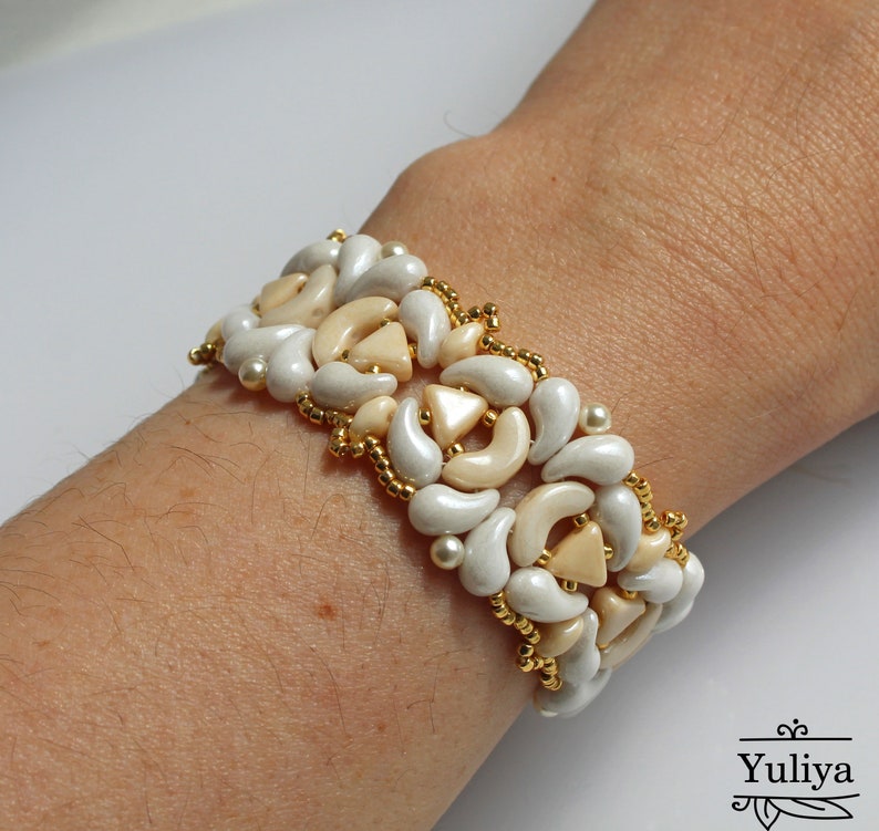 Op de afbeelding: Een witte en beige kralen armband met een geometrisch patroon. De armband is gemaakt met kleine, ronde kralen en grotere, traanvormige kralen.
