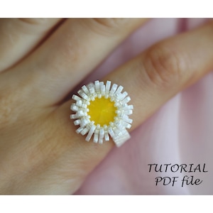 May include: A white beaded ring with a yellow center, resembling a daisy flower. The ring is worn on a finger and the image includes the text "TUTORIAL PDF file".