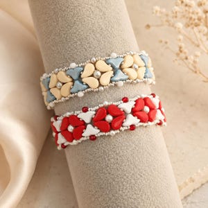 Könnte beinhalten: Zwei Perlenarmbänder mit einem sich wiederholenden Blumenmuster. Das obere Armband besteht aus cremefarbenen und hellblauen tropfenförmigen Perlen mit kleinen silbernen Akzenten. Das untere Armband zeigt rote und weiße Tropfenperlen mit roten Akzenten.