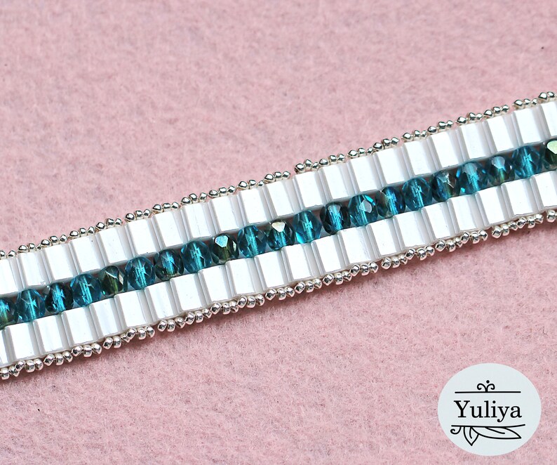 May include: A close-up of a white and teal beaded bracelet. The bracelet is made with small, square white beads and round teal beads. The beads are strung on a silver wire.