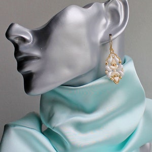 Op de afbeelding: Witte en gouden kralen oorbellen met een geometrisch ontwerp. De oorbellen zijn op een mannequin met een lichtblauwe sjaal.