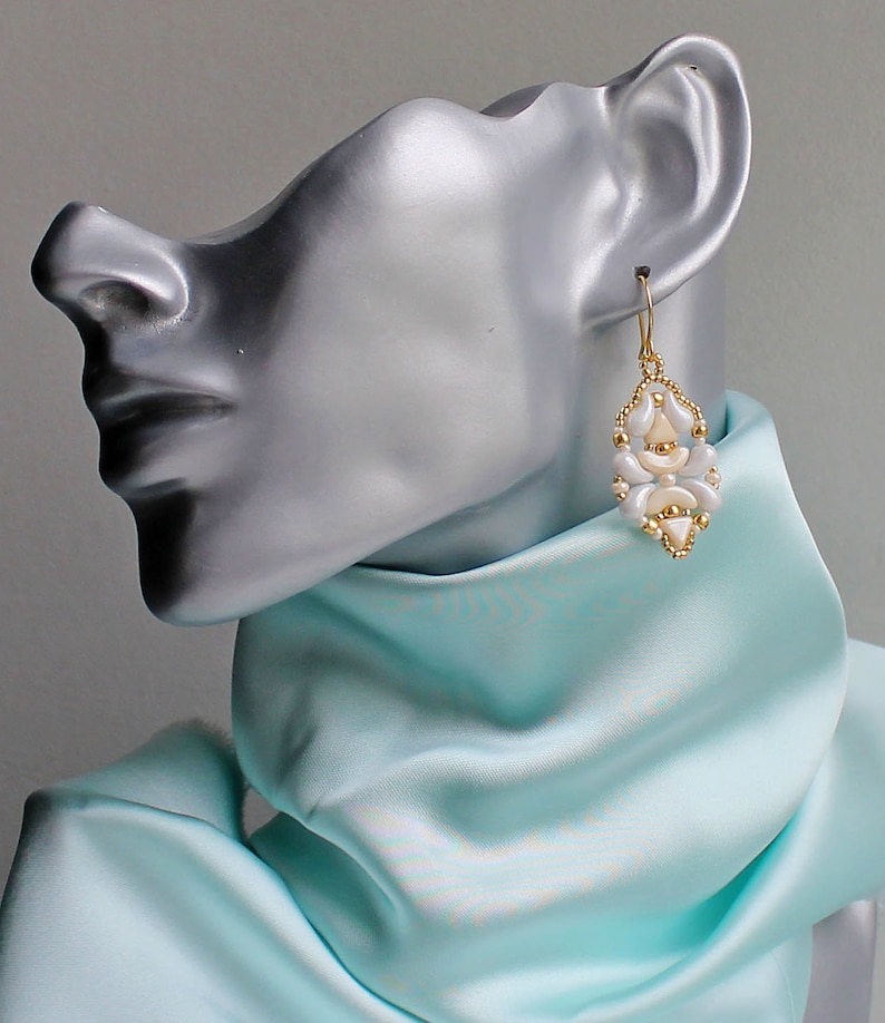 Op de afbeelding: Witte en gouden kralen oorbellen met een geometrisch ontwerp. De oorbellen zijn op een mannequin met een lichtblauwe sjaal.