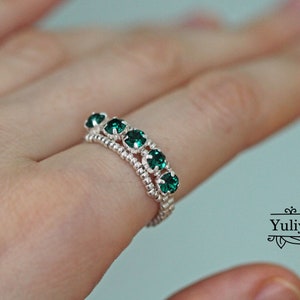 May include: A silver ring featuring a line of emerald green gemstones. The ring is worn on a finger.